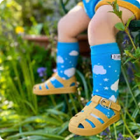 A child wears blue socks with a cloud design and sandals sitting on a bench amongst grassy area